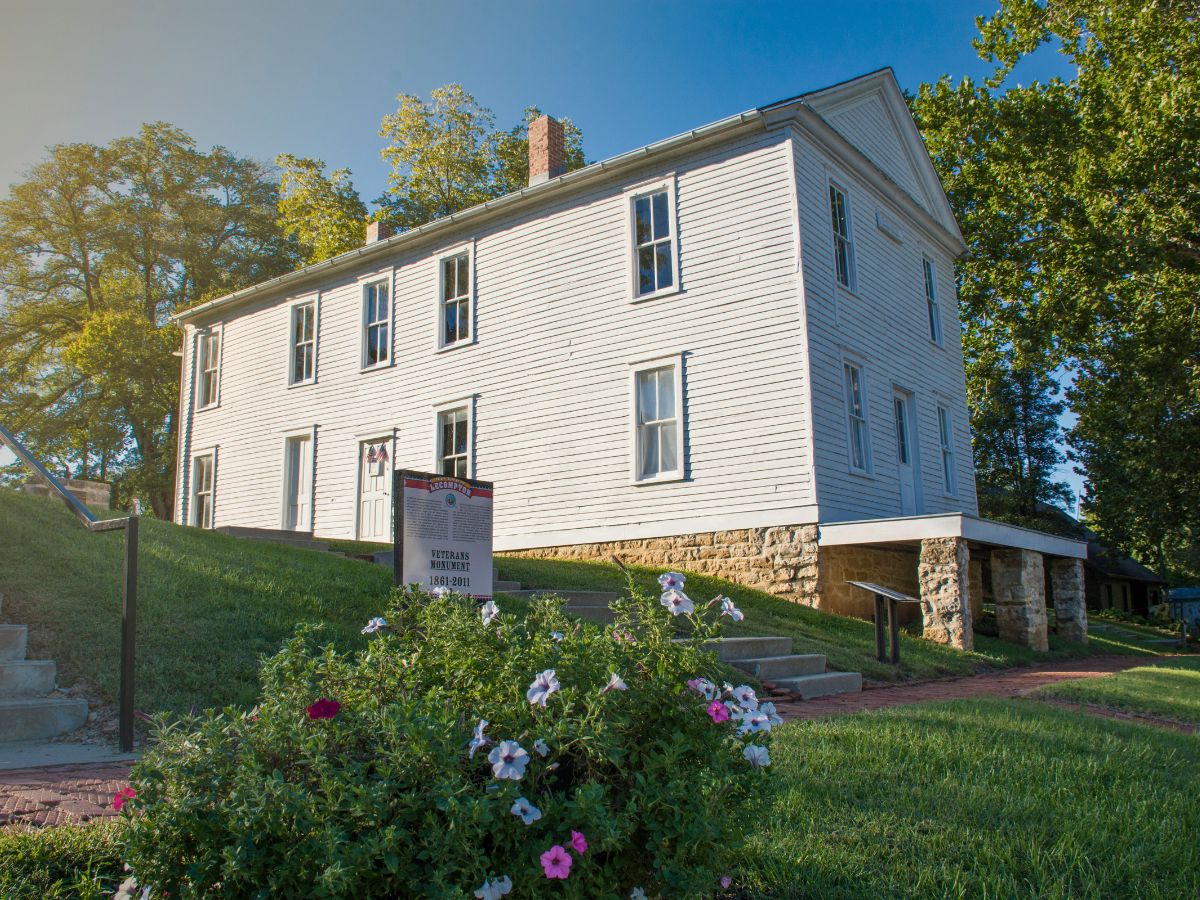 A white two-story house with blue shutters stands on a grassy lawn. A sign and flower bed are visible in the foreground. Trees surround the property.