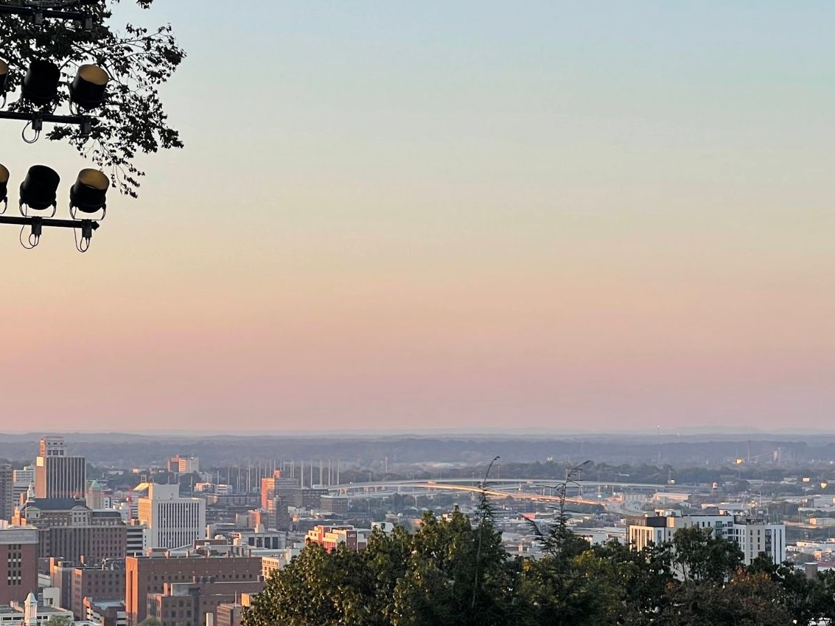 A cityscape at sunset with buildings and a bridge in the distance, seen from an elevated vantage point among trees. Stage lights are visible in the upper-left corner.