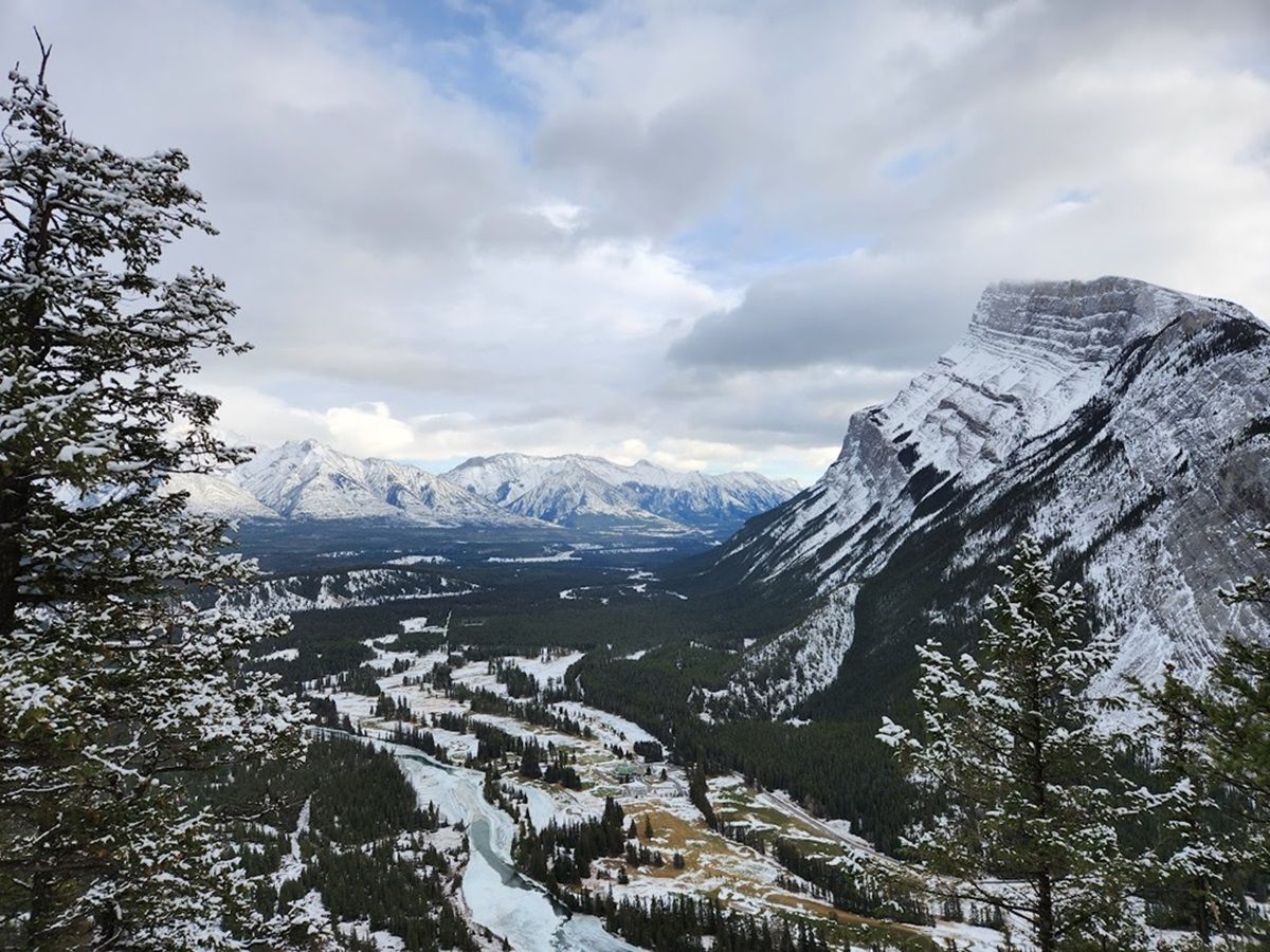 Snow-covered mountainous landscape with a large river valley flanked by evergreen trees. The sky above is partly cloudy, revealing patches of blue.