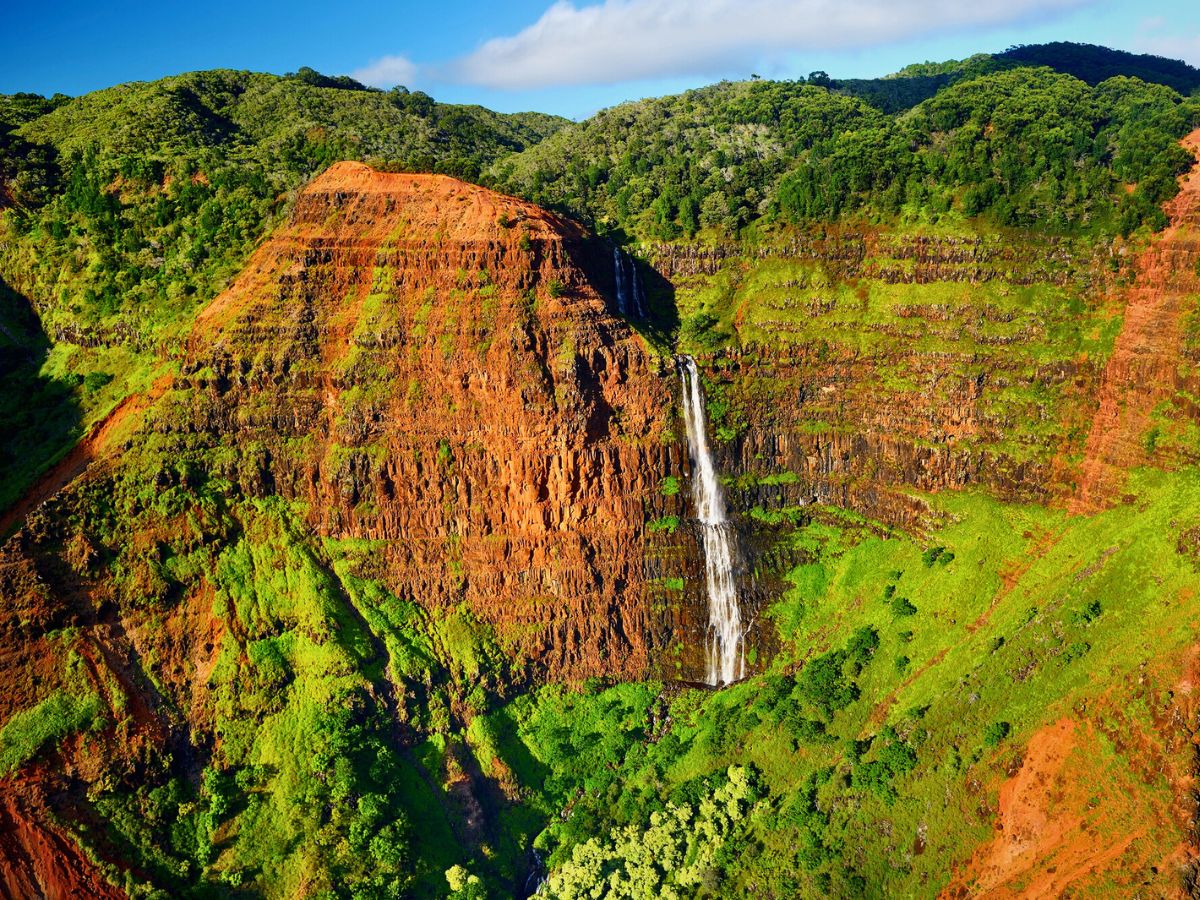 A waterfall cascades down a steep, rocky cliff surrounded by lush green vegetation under a clear blue sky.