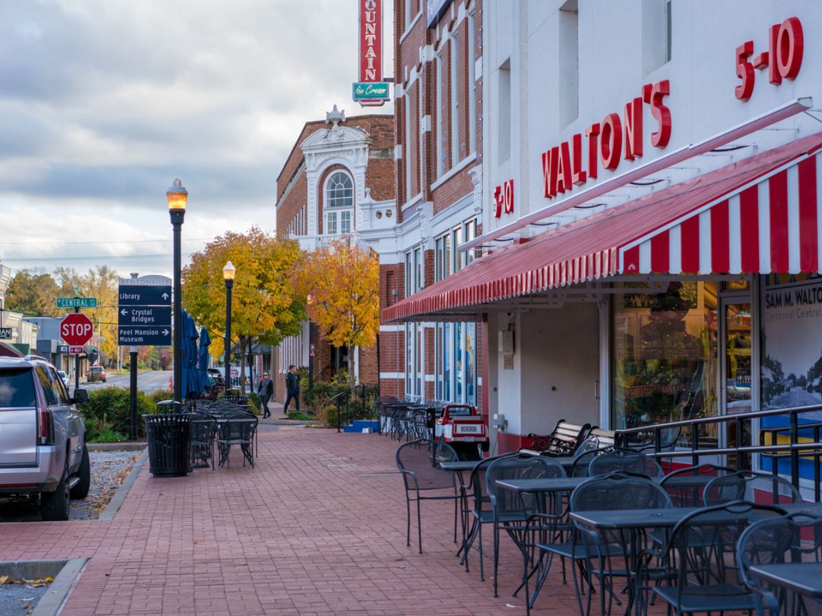 A sidewalk view of a historic district featuring storefronts, including a Walton's 5-10, outdoor seating, and autumn trees lining the brick street on a cloudy day.