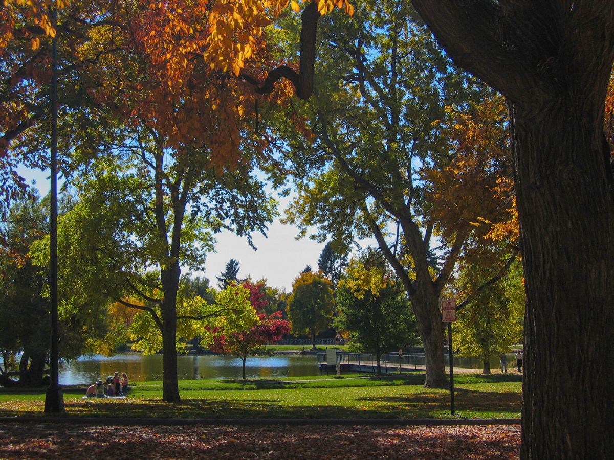 Park with large trees in autumn colors, a few people sitting by a lake, and a wooden dock in the background.
