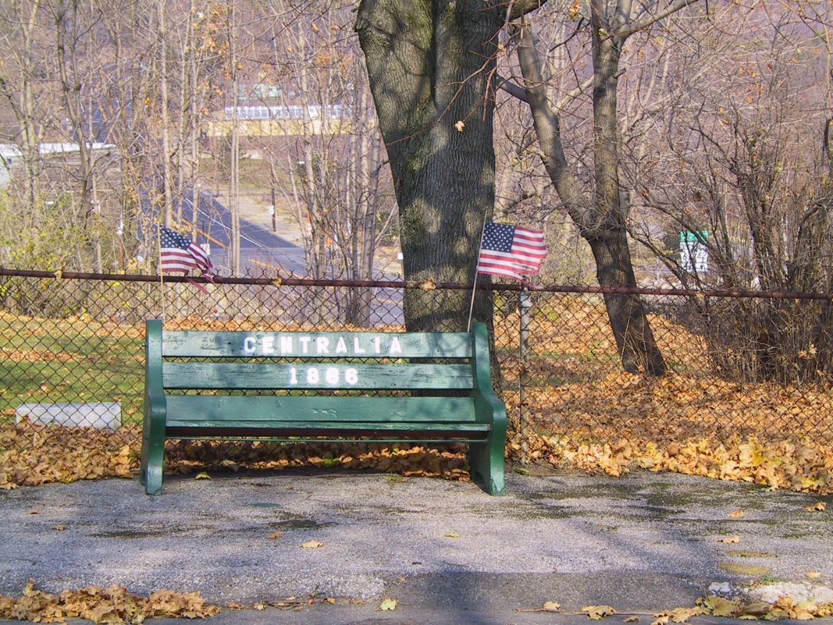 A green bench labeled "Centralia 1866" sits in a park with fallen leaves, two American flags, and a chain-link fence in the background.