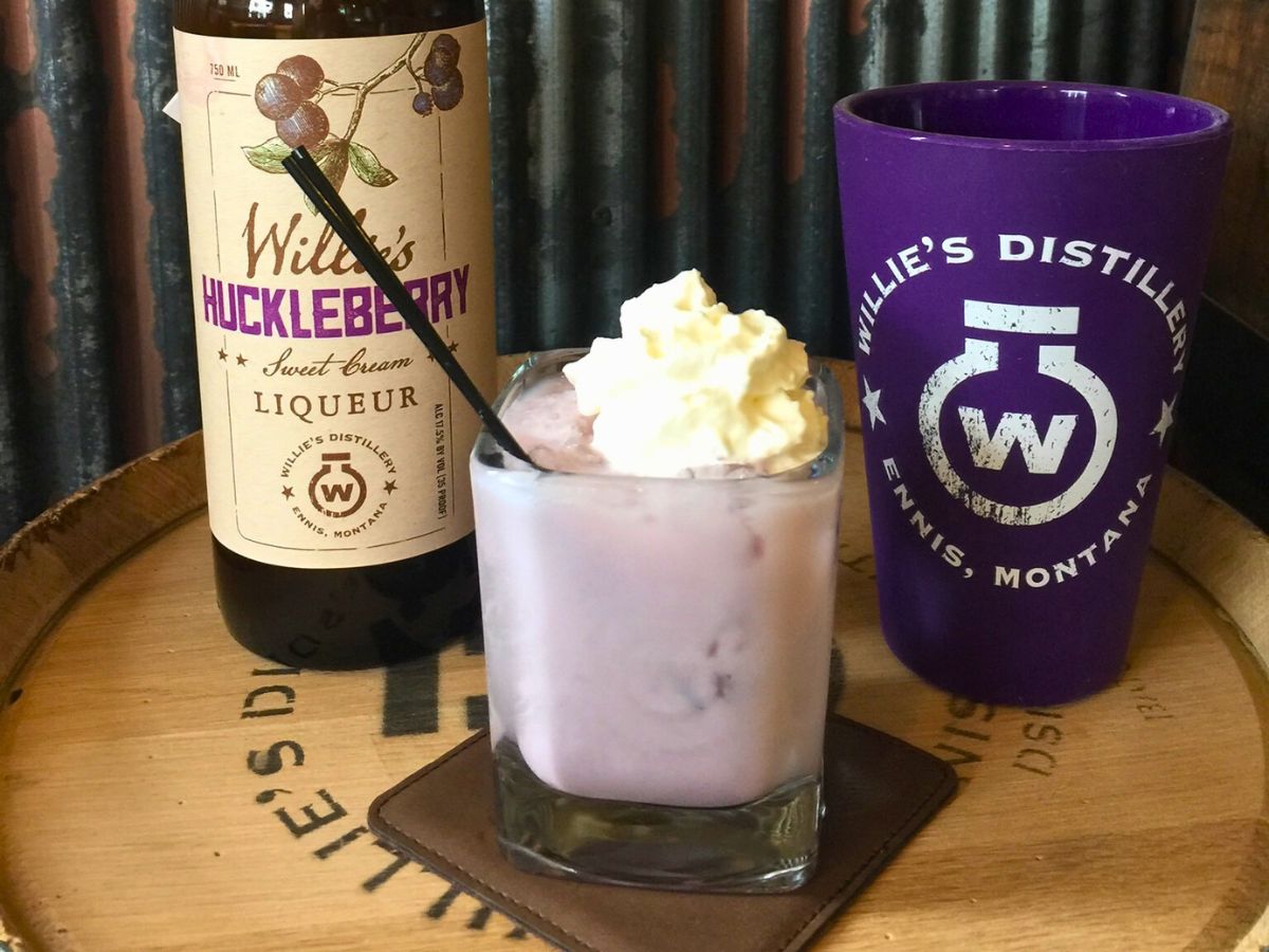 A glass of creamy purple cocktail topped with whipped cream, beside a bottle of wildberry huckleberry liqueur and a branded purple cup.