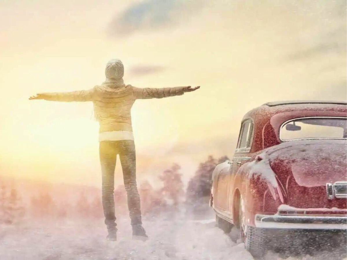 A person stands with arms outstretched in a snowy landscape near an old car, facing the sunrise or sunset.