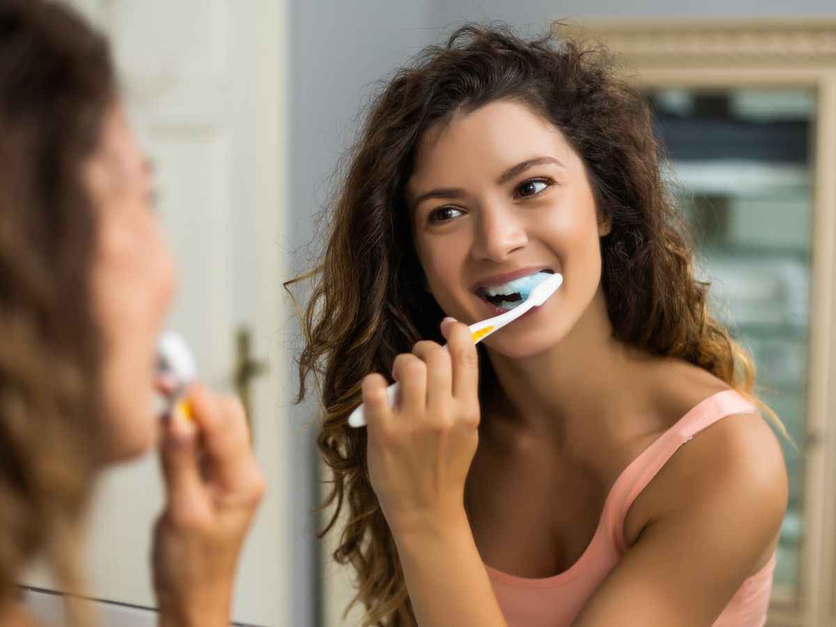 A woman with curly hair smiles while brushing her teeth in front of a mirror.