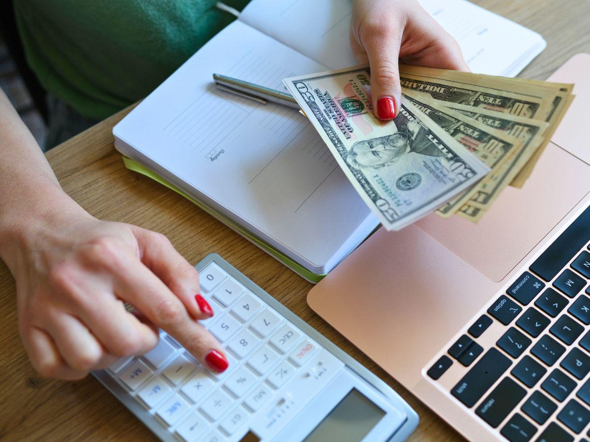 Person using a calculator and holding dollar bills next to an open notebook and a laptop on a wooden desk.