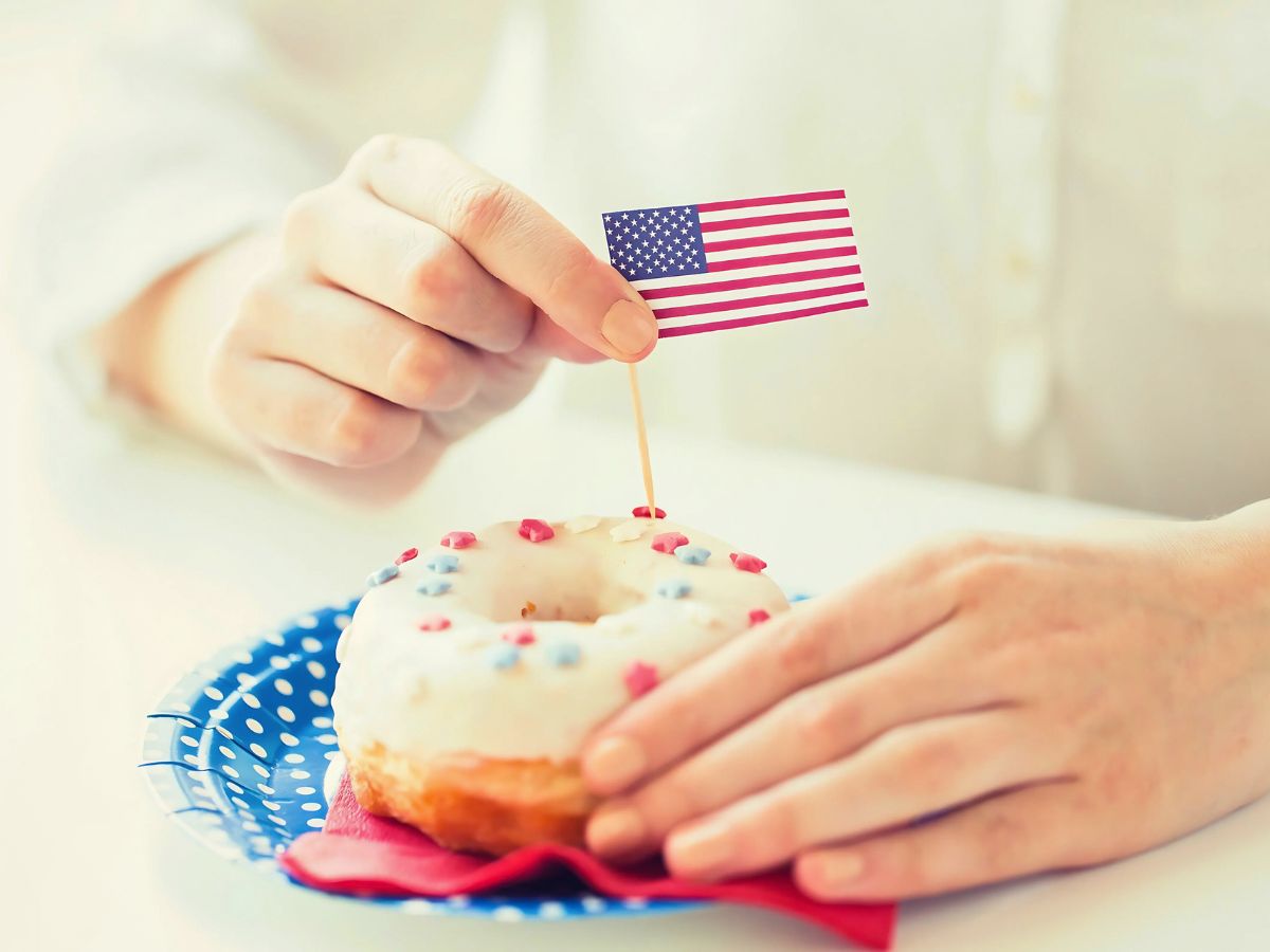 A woman places a small American flag on a white frosted donut with red and blue sprinkles, set on a polka dot plate.