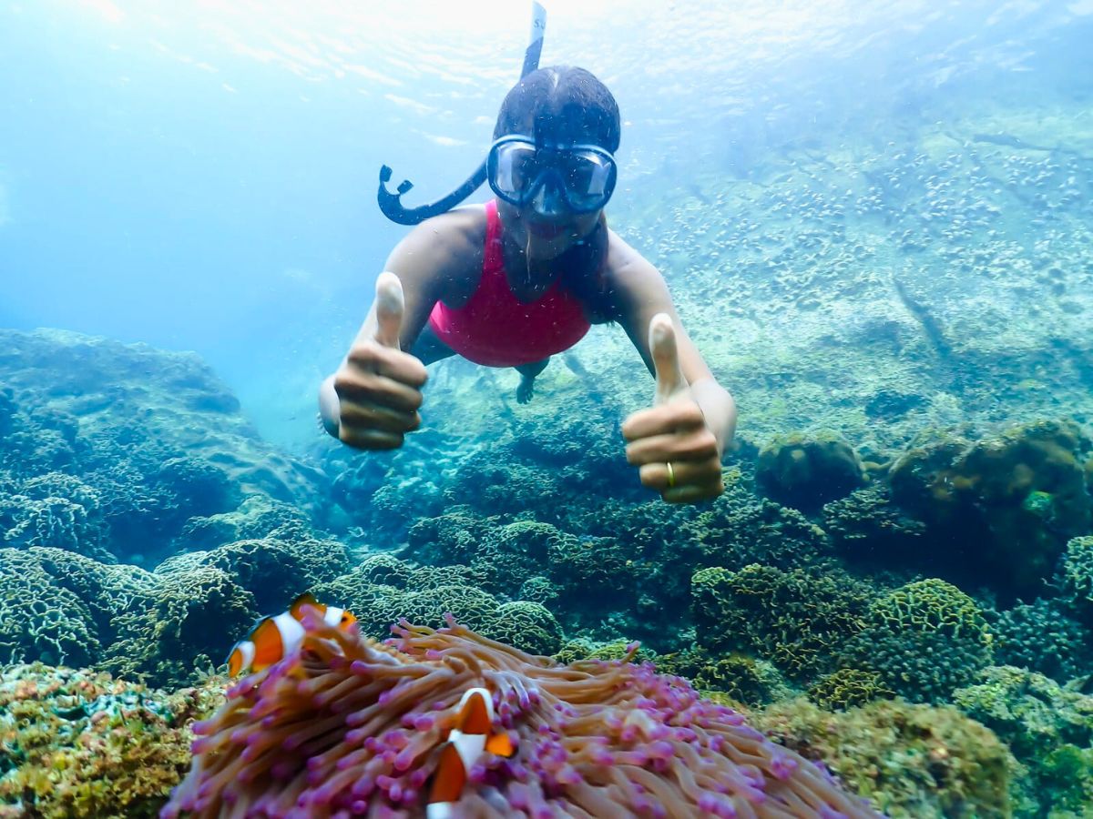 A person wearing snorkeling gear gives two thumbs up underwater near clownfish and sea anemone.