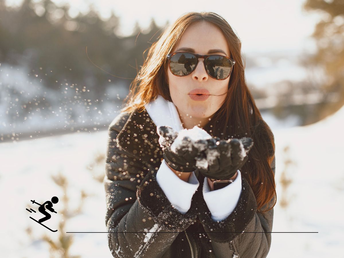 A woman wearing sunglasses blows snow from her hands in a snowy outdoor setting, with trees in the background. Icon of a skier is overlayed in the corner.