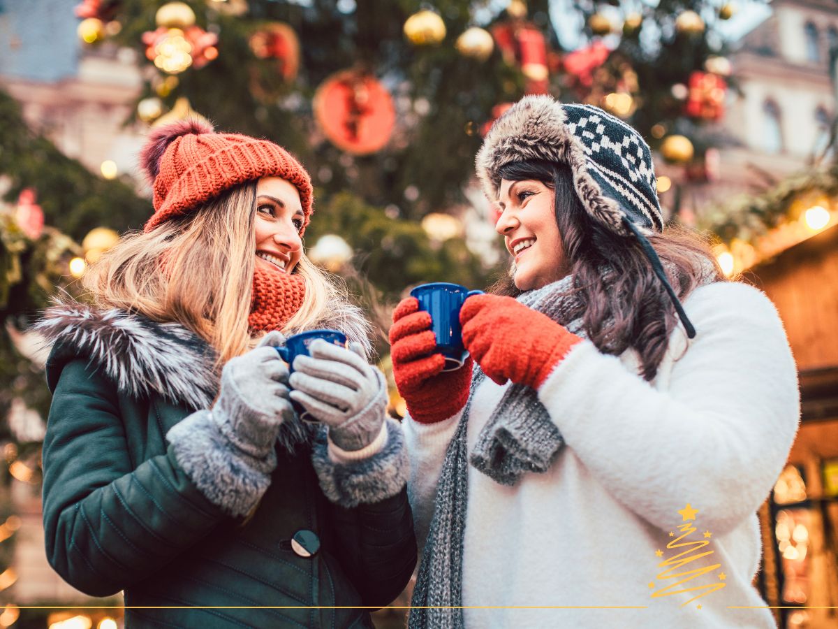Two people in winter clothing hold blue mugs, smiling at each other in front of a decorated Christmas tree.