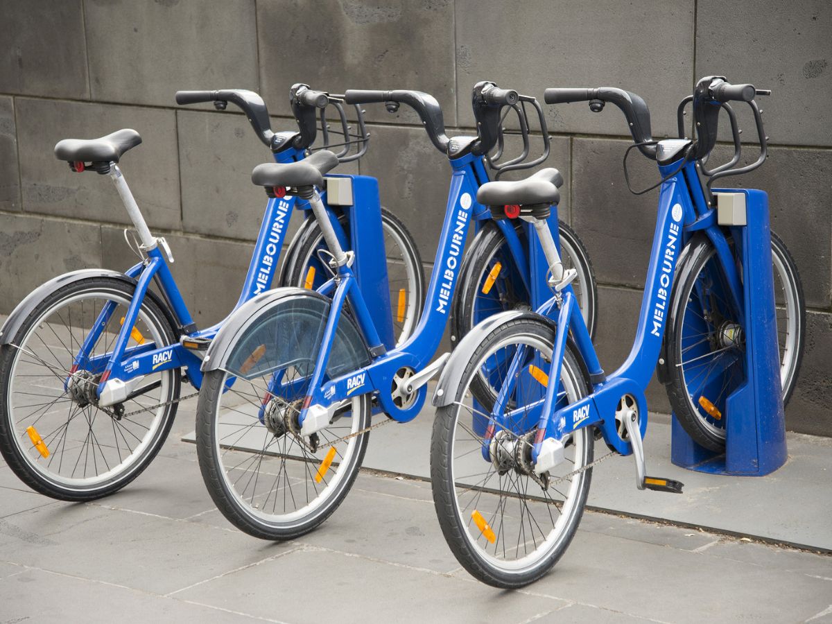 Four blue city bikes are parked at a bike-sharing station in an urban setting, adjacent to a grey concrete wall.