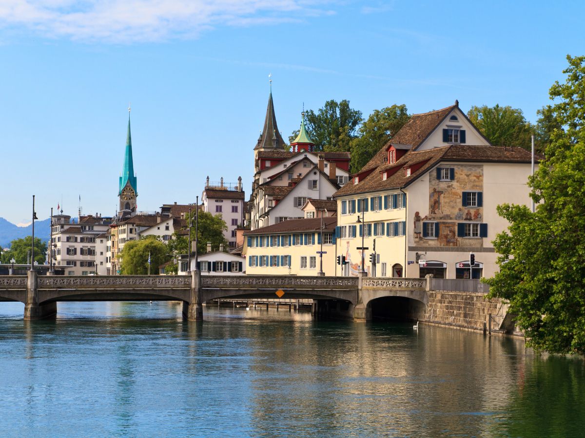 A scenic view of a historic bridge and old buildings along a river, with a church spire visible in the background under a clear blue sky.