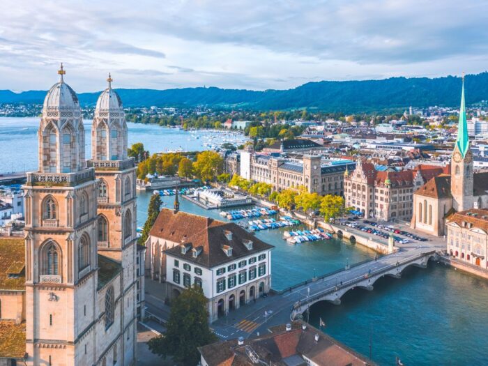 Aerial view of Zurich, showing the Grossmünster, the Limmat River, and surrounding cityscape with mountains in the background.