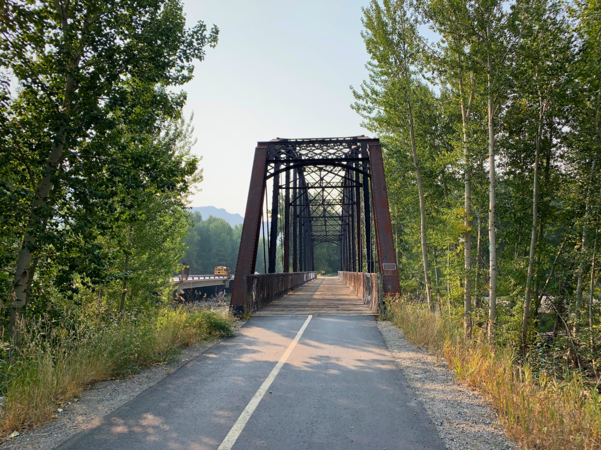 A paved path leads to a rusty iron truss bridge surrounded by tall trees under a clear sky.